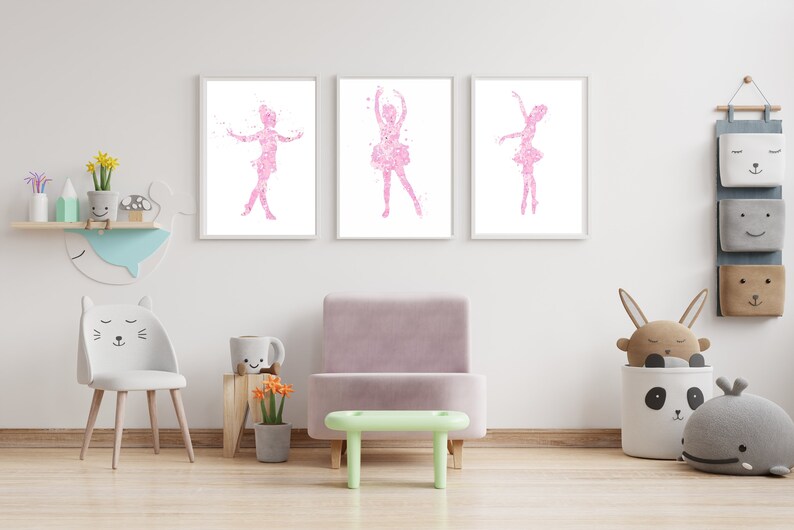 May include: Three framed prints of pink ballerina silhouettes on a white background. The prints are hanging on a white wall in a child's room. The room also features a light green stool, a white cat-shaped chair, and a gray storage bin with a panda face.