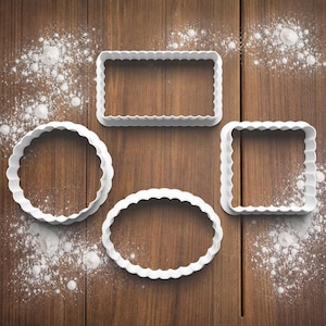 May include: Four white plastic cookie cutters in various shapes: a rectangle, a circle, a square, and an oval. Each cutter features a scalloped edge. Flour is scattered around the cutters on a wooden surface.