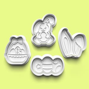 Puede incluir: Cuatro cortadores de galletas de plástico blanco con forma de diseños de Pascua: un conejito sosteniendo un huevo, una cesta de huevos, orejas de conejo y tres huevos.
