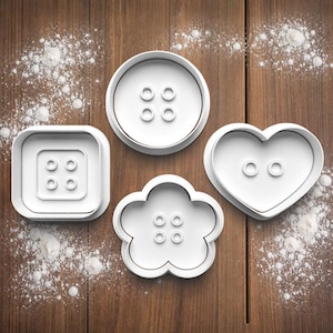 May include: Four white plastic cookie cutters shaped like buttons: a square, a circle, a flower, and a heart. Each cutter has button hole details. They are arranged on a wooden surface dusted with flour.
