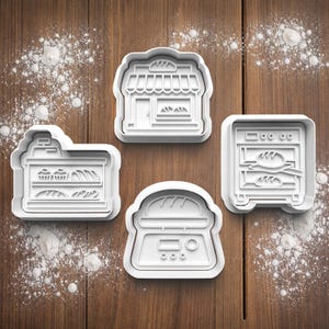May include: Four white plastic cookie cutters in the shapes of bakery items. The cutters depict a bakery shop, a display case, a bread scale, and an oven. The cutters are on a wooden surface with flour.