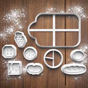 May include: A selection of white plastic cookie cutters in food shapes, such as a bunch of grapes, a sandwich, a cheese block, and a loaf of bread. The cutters are displayed on a wooden surface with scattered white powder.