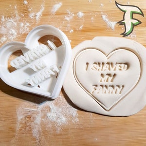 May include: White plastic heart-shaped cookie cutter with the text "I shaved my yinyas" and a white heart-shaped cookie with the text "I shaved my fanny" on a wooden surface.