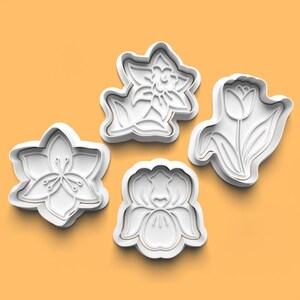 May include: Four white plastic cookie cutters shaped like different flowers. The cutters are on a light orange background.