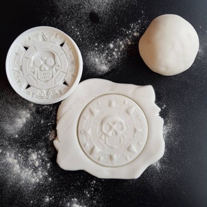 May include: White plastic cookie cutters shaped like a skull and crossbones. The skull has a detailed design and the crossbones are simple.