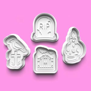 Może przedstawiać: Cztery białe wykrawacze do ciastek o tematyce Halloween. Kształty obejmują nagrobek z napisem "RIP", kruka na krzyżu, czaszkę ze świecą i bramę z nietoperzami. Tło jest różowe.