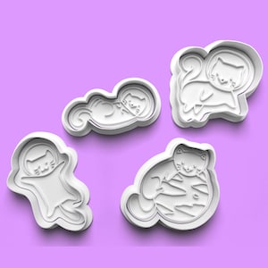 May include: Set of four white plastic cookie cutters shaped like cats in various poses. The cats are all wearing astronaut helmets and are in different positions, including sleeping, floating, and sitting on a planet.