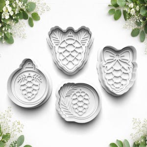 May include: Four white plastic cookie cutters in different shapes, including a round bauble, a pine cone, and a pine cone with a bow. The cutters are on a white surface, with greenery in the corners.