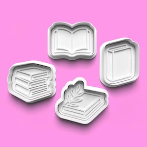 以下が含まれることがあります： 本型の白いプラスチック製クッキーカッター4つ。1つは開いた本、もう1つは閉じた本、2つは本の山です。カッターはピンク色の背景にあります。