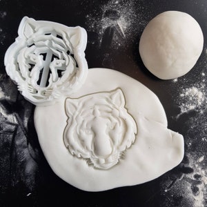 May include: White plastic cookie cutters in the shape of a tiger's head. The cutters are on a white surface with white dough.