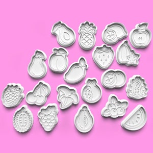 Op de afbeelding: Een set van 18 witte plastic koekjesvormen in de vorm van verschillende vruchten, waaronder een ananas, watermeloen, avocado, citroen en kersen. De vormen zijn gerangschikt op een roze achtergrond.