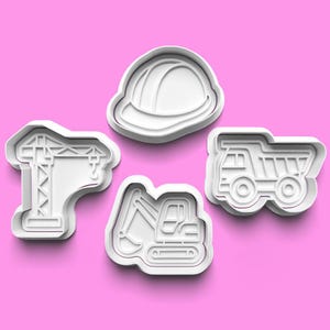 Può includere: Quattro formine per biscotti in plastica bianca a forma di veicoli e attrezzature edili. Le forme includono una gru, un casco, un escavatore e un dumper. Lo sfondo è di colore rosa.