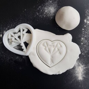 May include: A white heart-shaped cookie cutter with a geometric design and a white heart-shaped cookie dough imprint on a white surface.