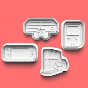 Może przedstawiać: Cztery białe plastikowe foremki do ciasteczek w kształcie konsol do gier wideo i kontrolerów. Formy znajdują się na różowym tle.