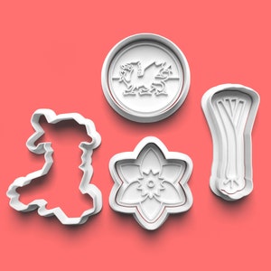 May include: A collection of white plastic cookie cutters in various shapes, including the outline of Wales, a daffodil, a leek, and a circular emblem with a dragon design. The cutters are set against a solid red background.