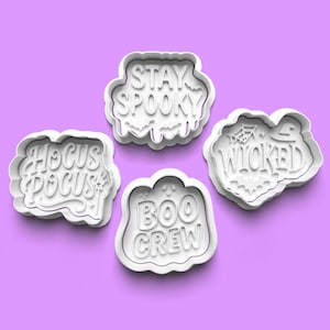 Puede incluir: Cuatro cortadores de galletas blancos de plástico con temática de Halloween sobre un fondo morado. Los cortadores tienen las frases: "Stay Spooky", "Hocus Pocus", "Boo Crew" y "Wicked". Cada cortador tiene un diseño y una fuente únicos.