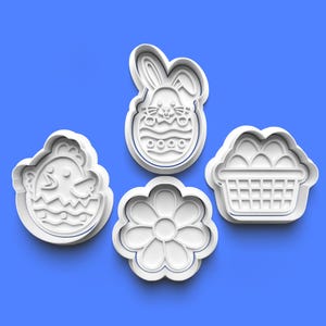 Puede incluir: Cuatro cortadores de galletas blancos de plástico con temática de Pascua. Las formas incluyen un pollito, un conejito en un huevo, una flor y una cesta de huevos. Los cortadores están dispuestos sobre un fondo azul.