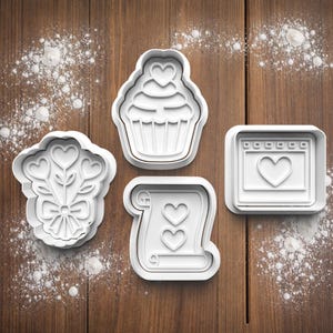 May include: Four white plastic cookie cutters in different shapes: a cupcake with a heart, a bouquet of hearts, a scroll with hearts, and a calendar with a heart. They are on a wooden surface with scattered white powder.