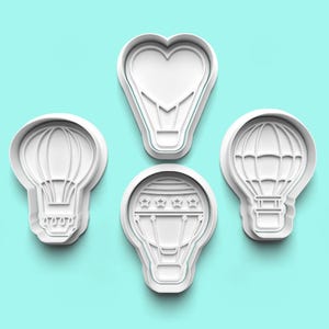 May include: Four white plastic cookie cutters shaped like hot air balloons. One cutter has a heart-shaped balloon, one has a balloon with a striped pattern, and two have balloons with a simple oval shape.
