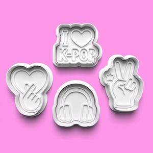 May include: Four white plastic cookie cutters on a pink background. The cutters are shaped like a heart with a hand gesture, headphones, a peace sign with a star, and the text "I Love K-Pop" with a heart.