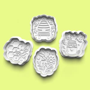 以下が含まれることがあります： ミツバチをテーマにしたデザインの、白いプラスチック製のクッキーカッター4つ。ハチの巣とミツバチ、ミツバチの巣箱、ハチミツの瓶とハニーディッパー、花とミツバチの形があります。カッターは明るい緑色の背景にあります。