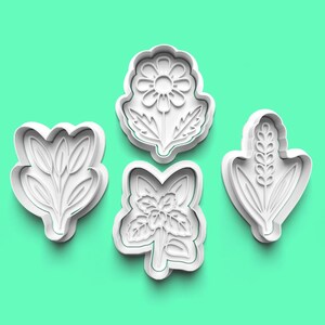 May include: Four white plastic floral-shaped cookie cutters. The shapes include a daisy, a sprig of leaves, a cluster of flowers, and a wheat stalk. The cutters are on a light green background.