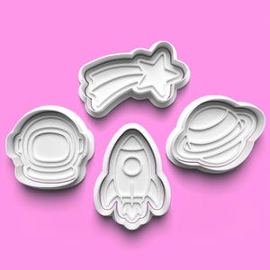 Puede incluir: Cuatro cortadores de galletas de plástico blanco con formas de temática espacial: un casco de astronauta, un cohete, una estrella fugaz y un planeta con anillos. Los cortadores están dispuestos sobre un fondo rosa.