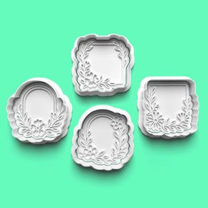 May include: Four white cookie cutters in different shapes: a square, a rectangle, and two oval shapes. Each cutter has a floral and leaf design around the edges. The cutters are arranged on a solid teal background.