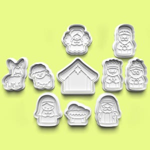 Op de afbeelding: Een verzameling witte plastic koekjessnijders in verschillende vormen, waaronder een engel, een ezel, een schaap, een stal en figuren die de kerststal voorstellen. De snijders zijn gerangschikt op een lichtgroene achtergrond, ideaal voor bakken.