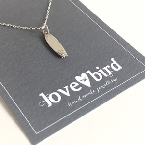 Op de afbeelding: Zilveren ketting met een hanger in de vorm van een surfboard. De hanger heeft een klein vogelontwerp. De ketting ligt op een grijze kaart met de tekst "lovebird hand-made jewellery".