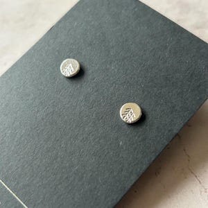 May include: A pair of silver stud earrings featuring a tree design. The earrings are displayed on a black background.