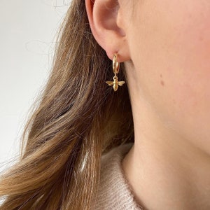 May include: A gold-colored hoop earring with a small, detailed bee charm. The bee is suspended from the hoop, adding a touch of nature-inspired elegance. The earring is worn on a person's ear, showcasing its design and style.