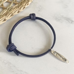 Op de afbeelding: Een donkerblauwe leren armband met een zilveren bedel in de vorm van een surfboard. De bedel heeft een gedetailleerd ontwerp van een surfboard met een peddel.