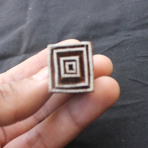 May include: A wooden block printing stamp with a square design featuring concentric squares. The stamp is brown with white lines outlining the squares.