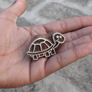 Puede incluir: Un sello de madera con forma de tortuga con un contorno blanco. El sello se sostiene en la mano de una persona.