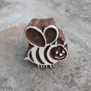 May include: A wooden block stamp with a bee design. The bee is carved in white and has a black and white striped body. The bee has a smiling face.