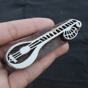 May include: A wooden block printing stamp with a detailed design of a sitar, a traditional Indian musical instrument. The stamp is brown with white details.