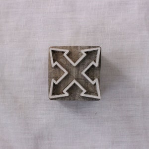 Op de afbeelding: Een houten blokstempel met een wit ontwerp van vier pijlen die in verschillende richtingen wijzen. De stempel is bruin en heeft een vierkante vorm.