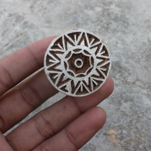 May include: A white wooden block printing stamp with a detailed geometric design. The design features a central circle with a smaller circle inside, surrounded by a ring of intricate patterns.