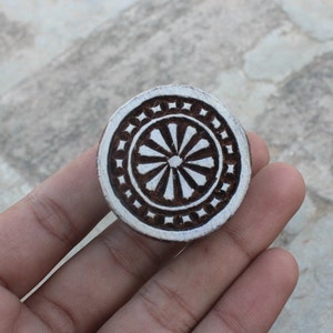 May include: A round wooden block printing stamp with a white background and a brown design. The design is a circular pattern with a flower in the center.