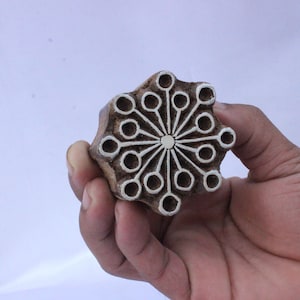 May include: A wooden block printing stamp with a white design of a sunburst with a circle in the center. The stamp is brown and has a rough texture.