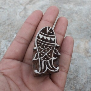 以下が含まれることがあります： 魚のデザインが細かく刻まれた木製の版画スタンプ。魚は茶色の背景に白く彫られています。