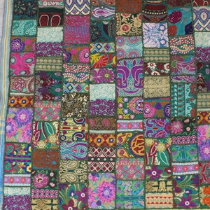 Op de afbeelding: Een kleurrijk patchwork textiel met een verscheidenheid aan patronen. De vierkanten hebben bloemen-, paisley- en geometrische ontwerpen in roze, paars, groen en goud. Een gele rand en een blauw gestreepte rand zijn zichtbaar.