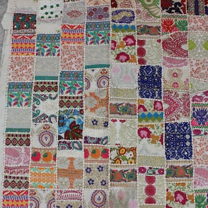 May include: A patchwork textile featuring a variety of colorful, patterned squares. The squares display floral, paisley, and geometric designs in shades of pink, blue, green, and orange. The edges are stitched together with a cream-colored thread.