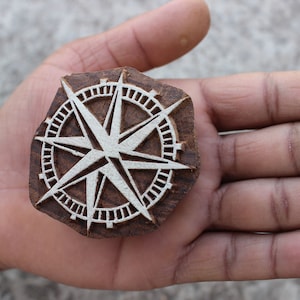 Può includere: Un timbro da stampa in legno con un design a stella bianca all'interno di un cerchio. Il timbro è intagliato in legno marrone scuro.