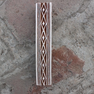 Op de afbeelding: Een houten drukblok met een wit en bruin geometrisch patroon. Het patroon bestaat uit herhalende ruiten.