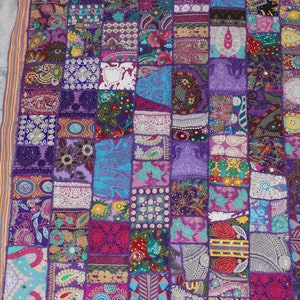 May include: A colorful patchwork wall hanging with a purple background. The hanging is made of various fabrics and textures, including velvet, cotton, and embroidery. It features a variety of patterns and designs, including floral motifs, paisley patterns, and geometric shapes. The hanging is adorned with beads and sequins, adding a touch of sparkle and dimension.