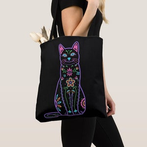 Puede incluir: Bolsa de tela negra con un dise&ntilde;o floral colorido de un gato. El gato est&aacute; contorneado en morado y tiene flores rosas, azules y verdes en su cuerpo.