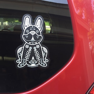 May include: Black and white sticker of a French bulldog with a Day of the Dead design. The sticker is on the back window of a red car.