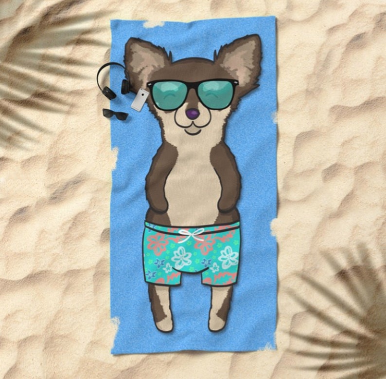 May include: A brown and white chihuahua wearing sunglasses and blue and green floral swim trunks is lying on a blue beach towel. The towel is on a sandy beach.